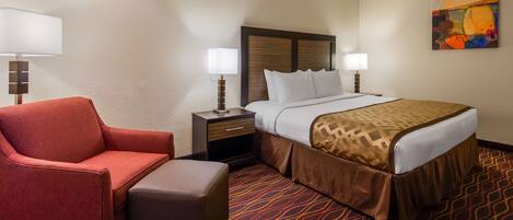 A hotel room with a bed, a red armchair, a nightstand, and a lamp.