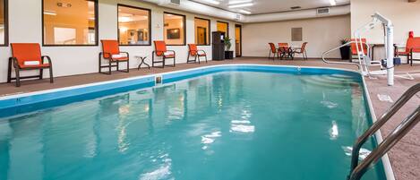 An indoor swimming pool with clear blue water, surrounded by orange chairs and a poolside table.