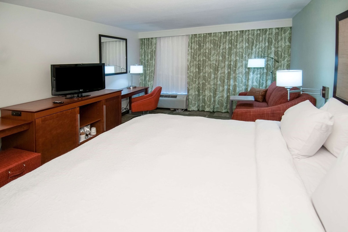 A hotel room with a large bed, a desk with a television, a chair, and a window with curtains.