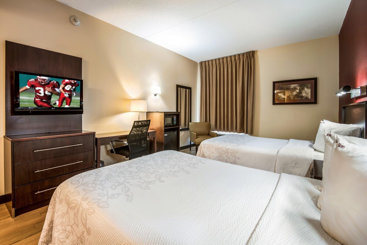 A hotel room with two beds, a desk with a chair, a television, a lamp, and a painting on the wall.