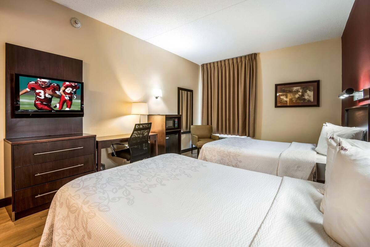 A hotel room with two beds, a desk with a chair, a television, a lamp, and a painting on the wall.