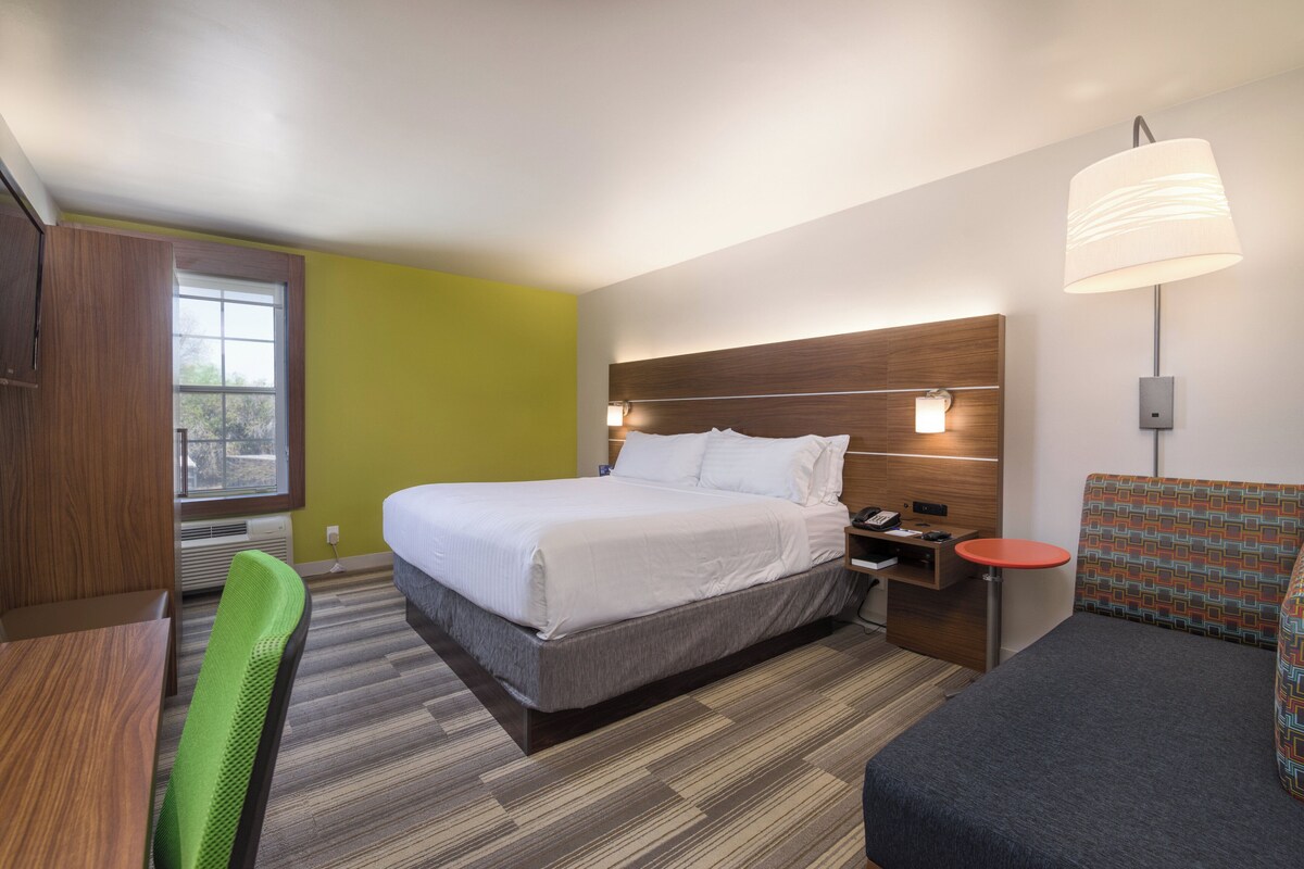 A hotel room with a bed, a desk, a chair, a nightstand, a lamp, and a window.
