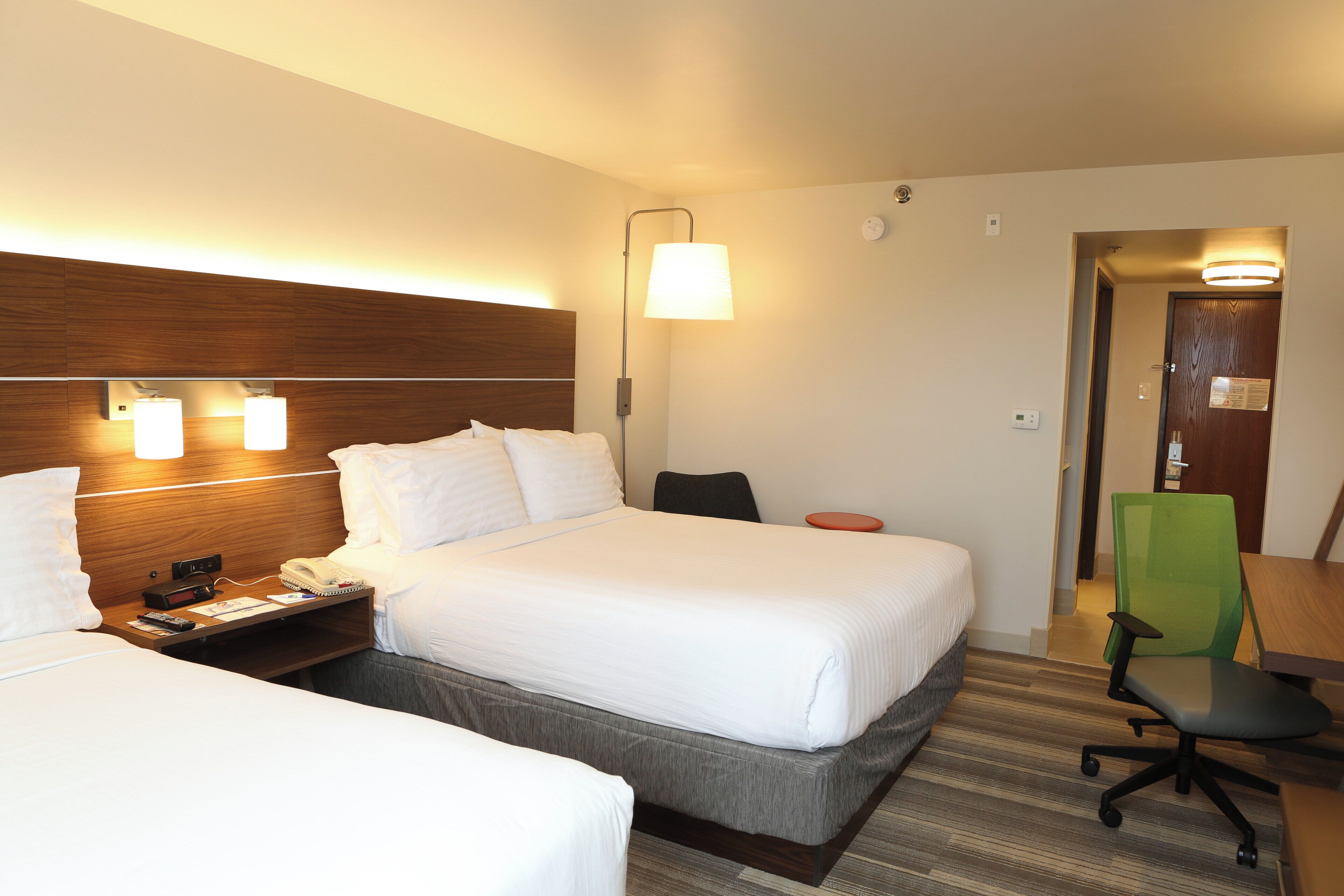 A hotel room with two beds, a desk, a chair, a lamp, and a door.
