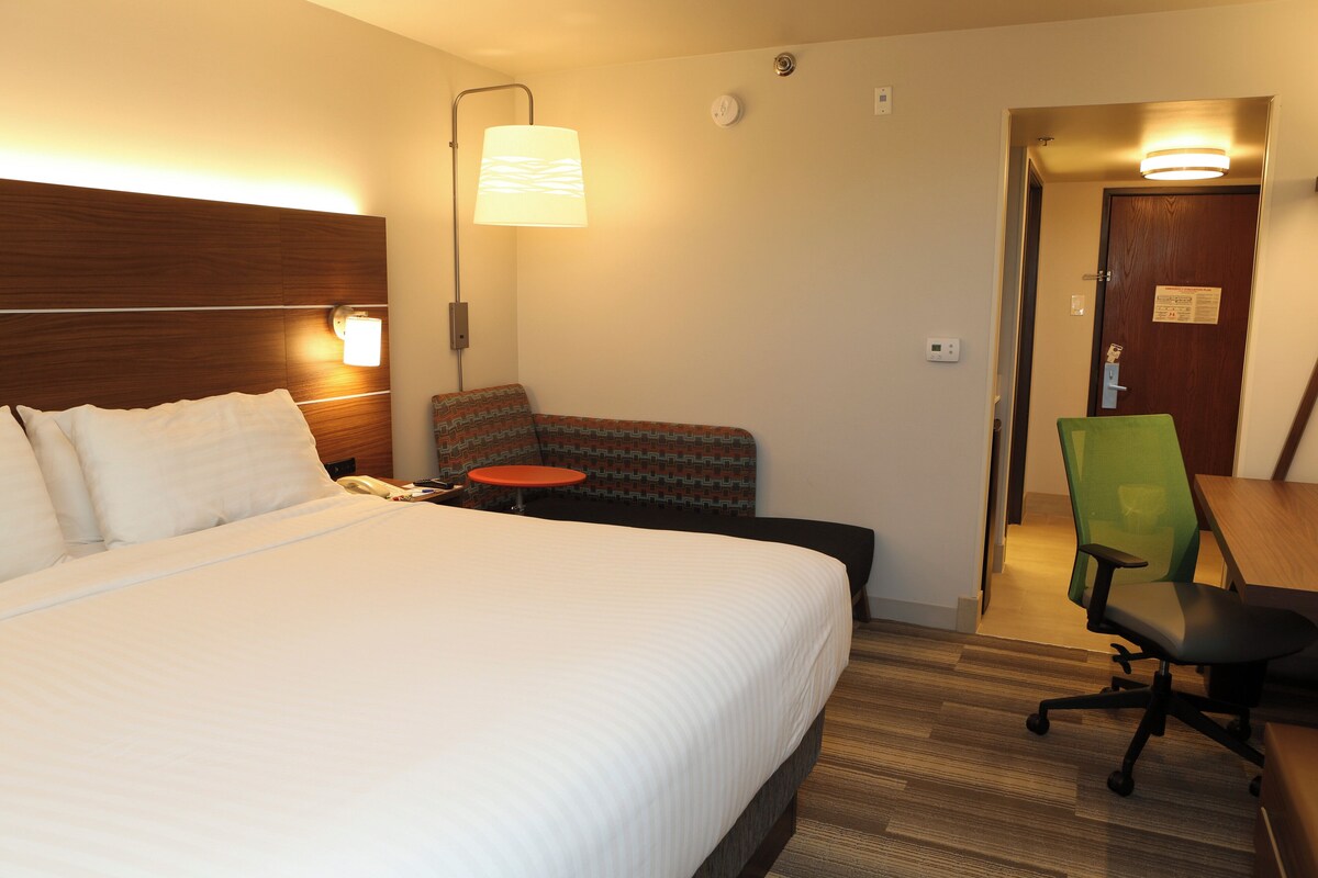 A hotel room with a bed, bedside lamp, chair, desk, and a door leading to another room.