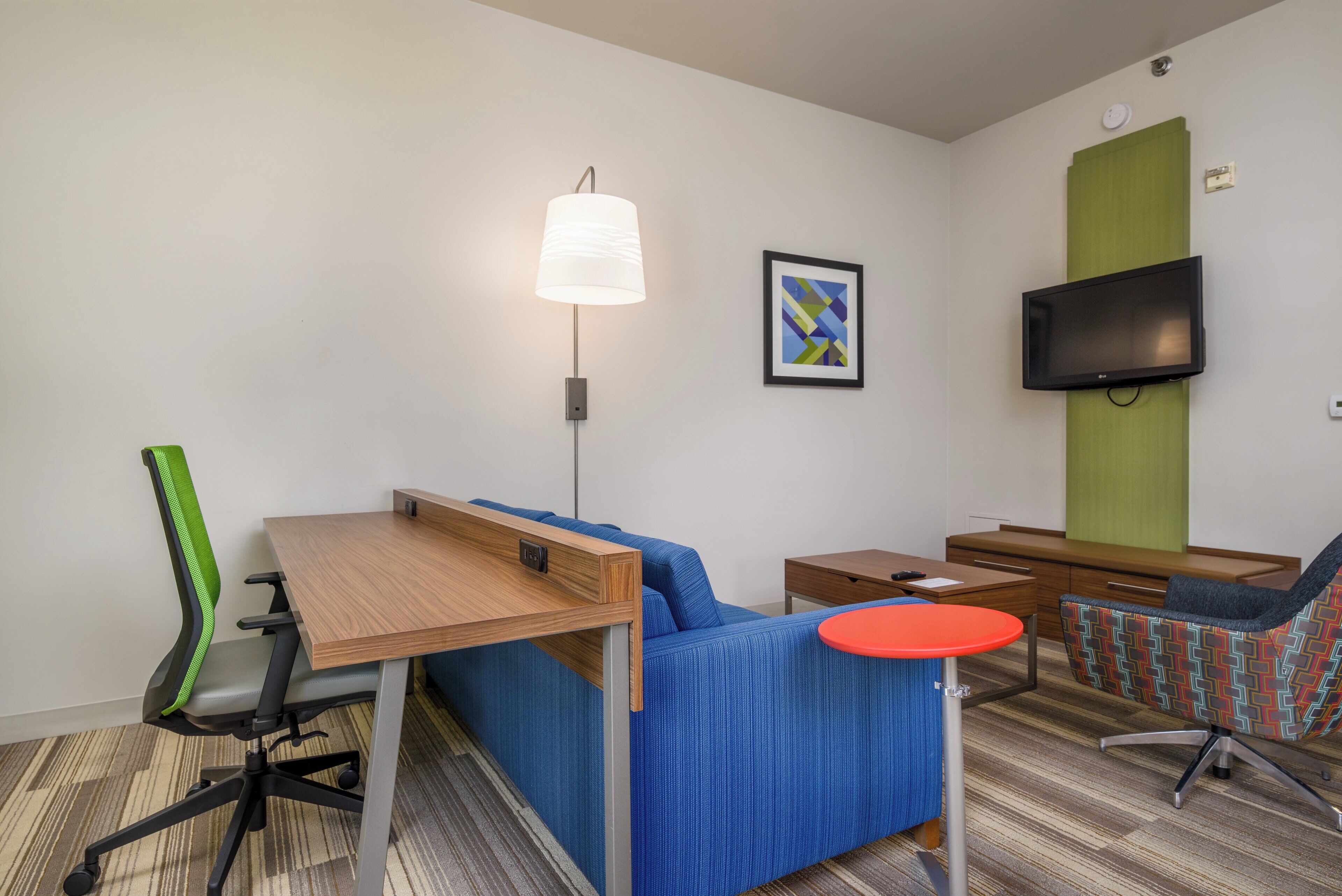 A hotel room with a sofa, a desk, a chair, a TV, and a lamp.