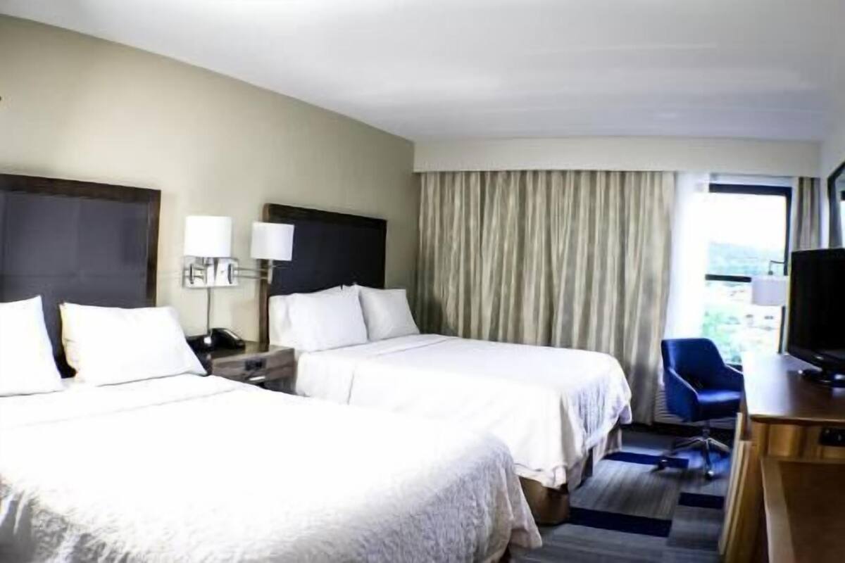 A hotel room with two beds, a desk with a computer, a chair, and a window with curtains.