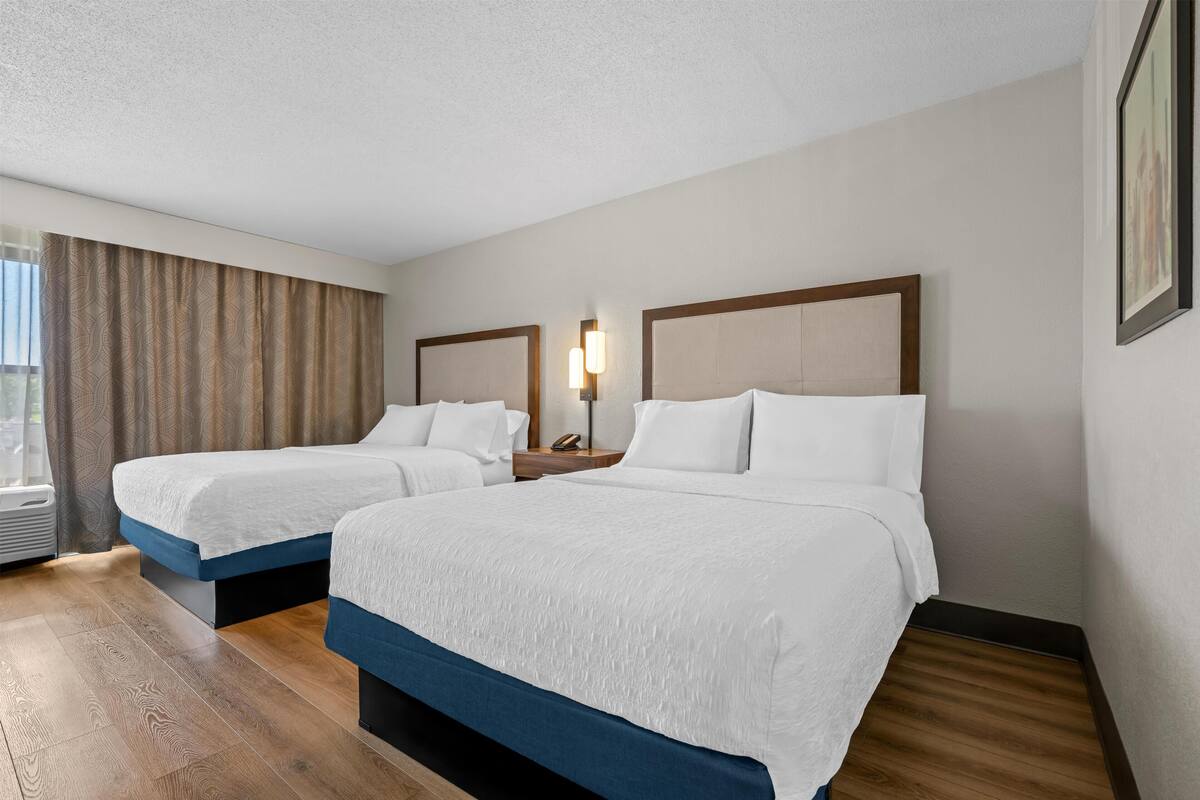 A hotel room with two beds, a bedside table with a lamp, a wall-mounted picture, and a window with curtains.