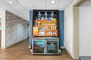 A vending machine with snacks and beverages, located in a hallway with patterned walls and wooden flooring. - Hampton Inn Birmingham/Trussville (Birmingham)