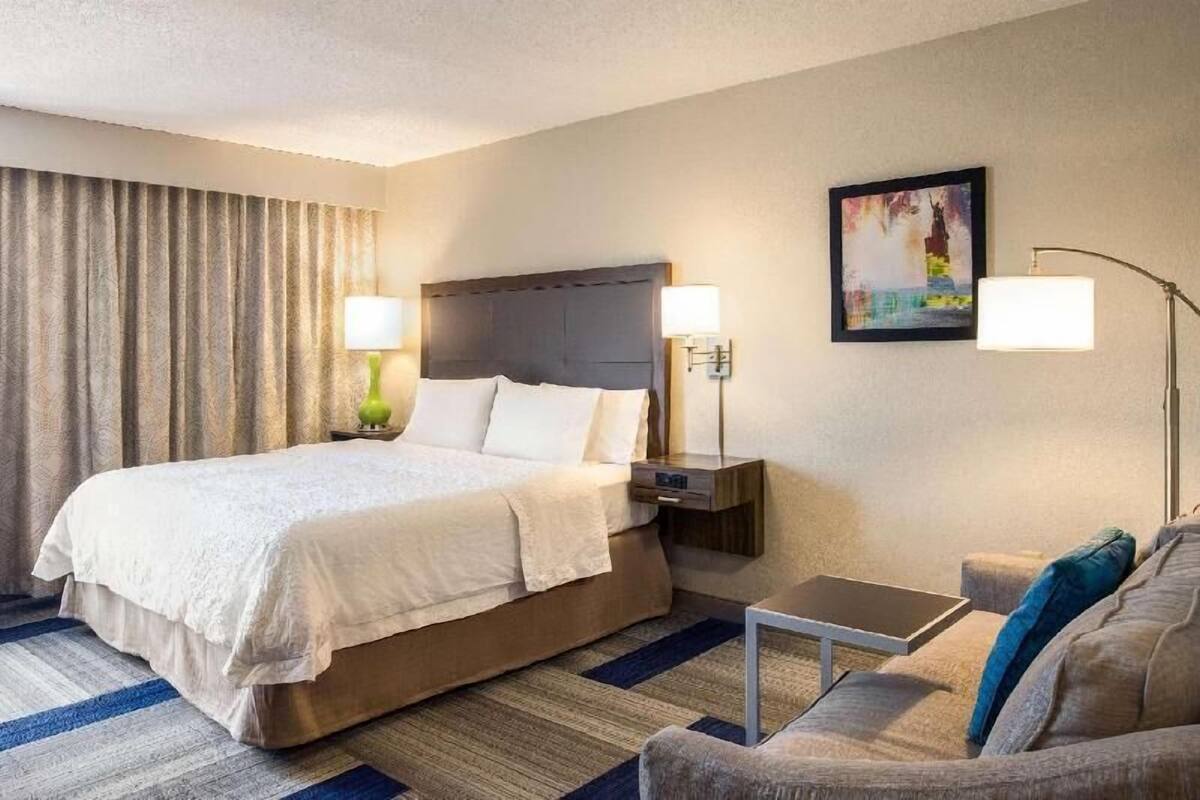 A hotel room with a large bed, bedside tables with lamps, a small table, a sofa, and a painting on the wall.