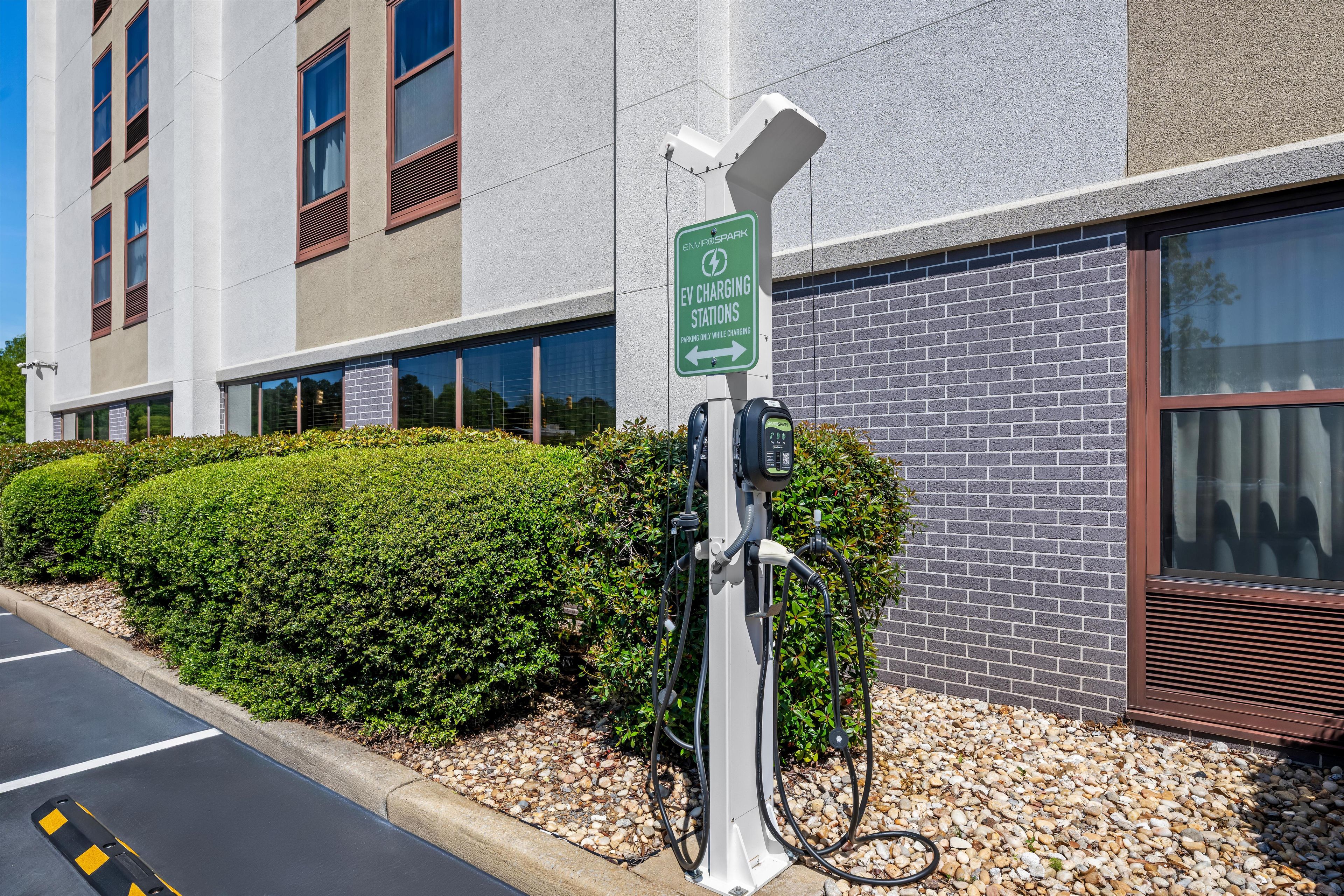An EV charging station with a sign indicating it is for EV charging, a building with large windows, and a parking area with a gravel path.