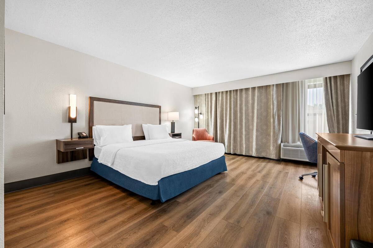 A hotel room with a large bed, a desk, a chair, a TV, and a window with curtains.