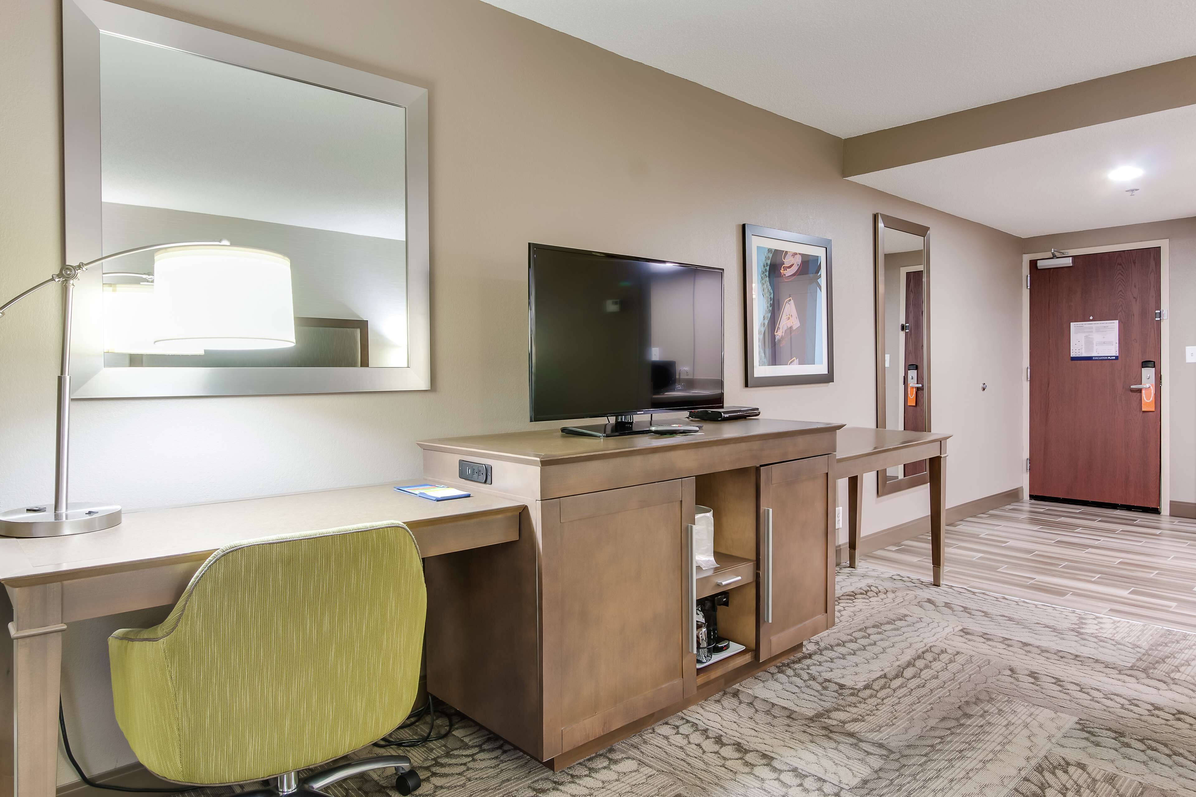 A hotel room with a desk, chair, TV, and a large mirror.