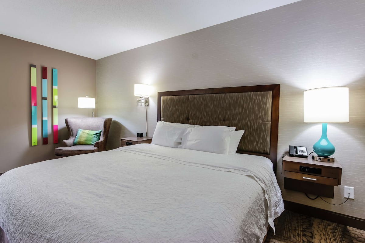 A hotel room with a large bed, a nightstand with a lamp, a chair, and a wall-mounted lamp.