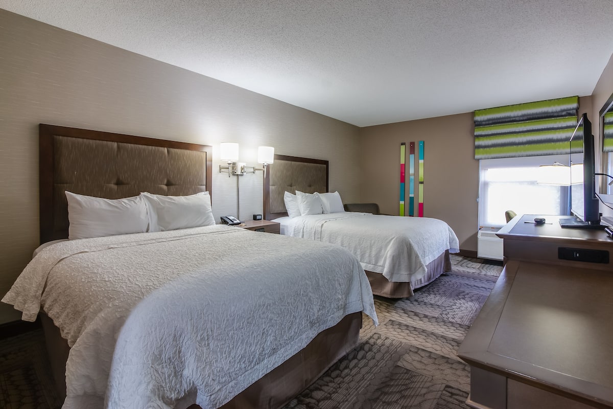 A hotel room with two beds, a desk, a chair, a lamp, and a window with curtains.