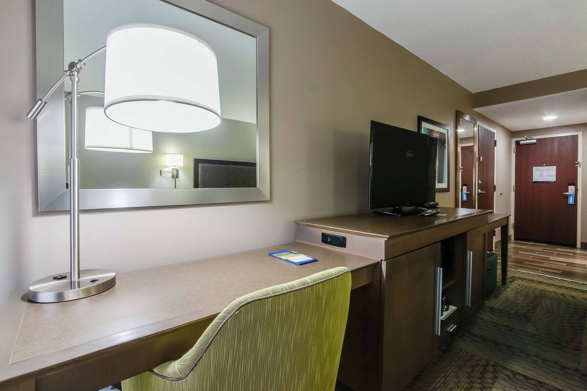 A hotel room with a desk, a chair, a large mirror, a television, and a door.