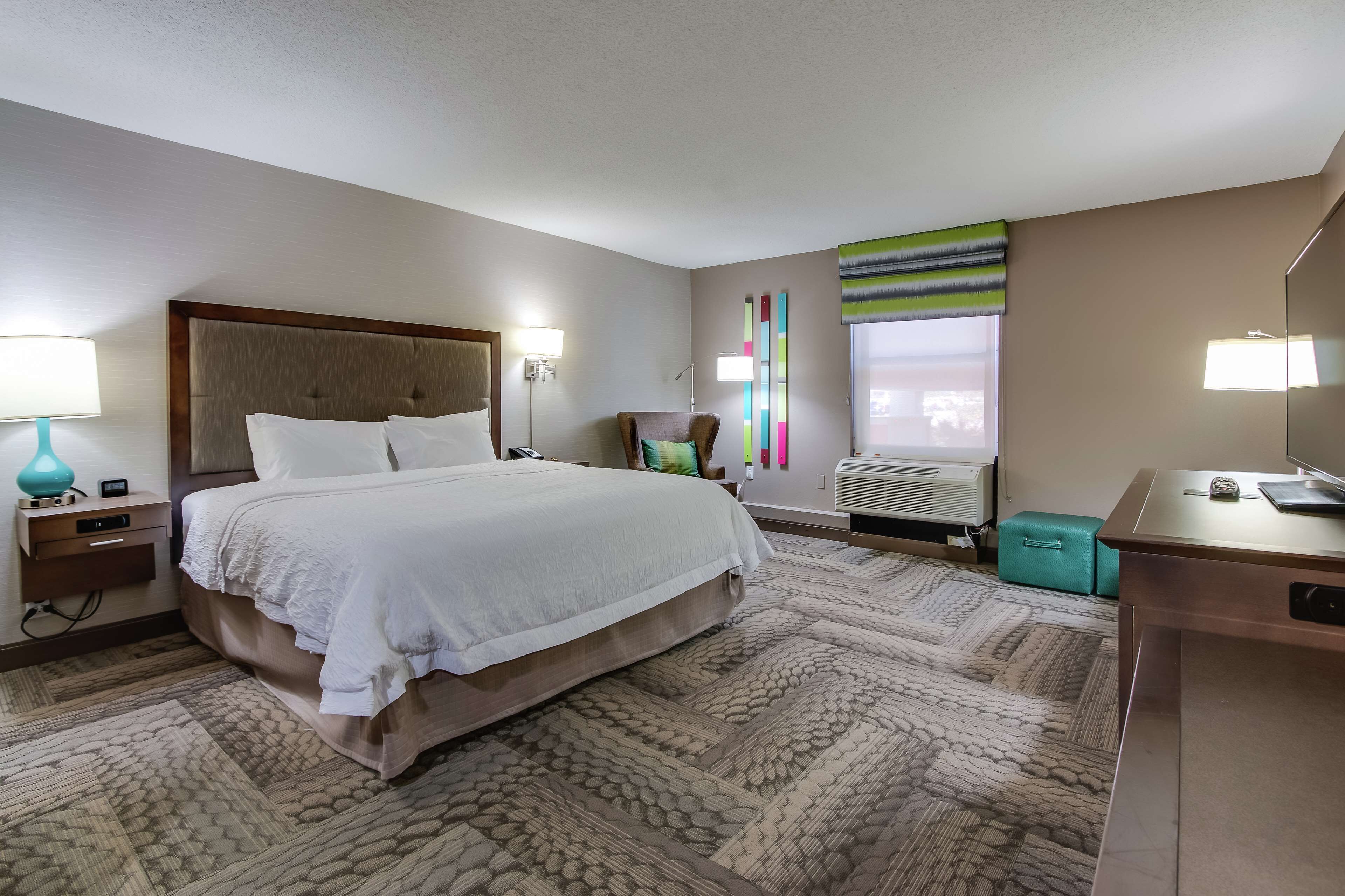 Una habitación de hotel con una cama grande, un escritorio, una silla, una lámpara y un ventanal con persianas.