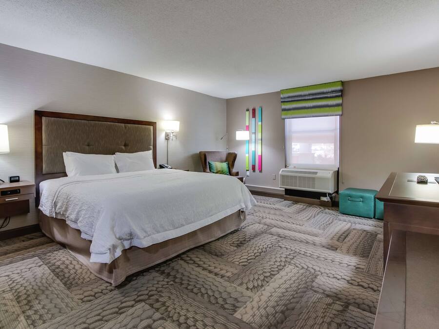 A hotel room with a large bed, a desk, a chair, a lamp, and a window with blinds.