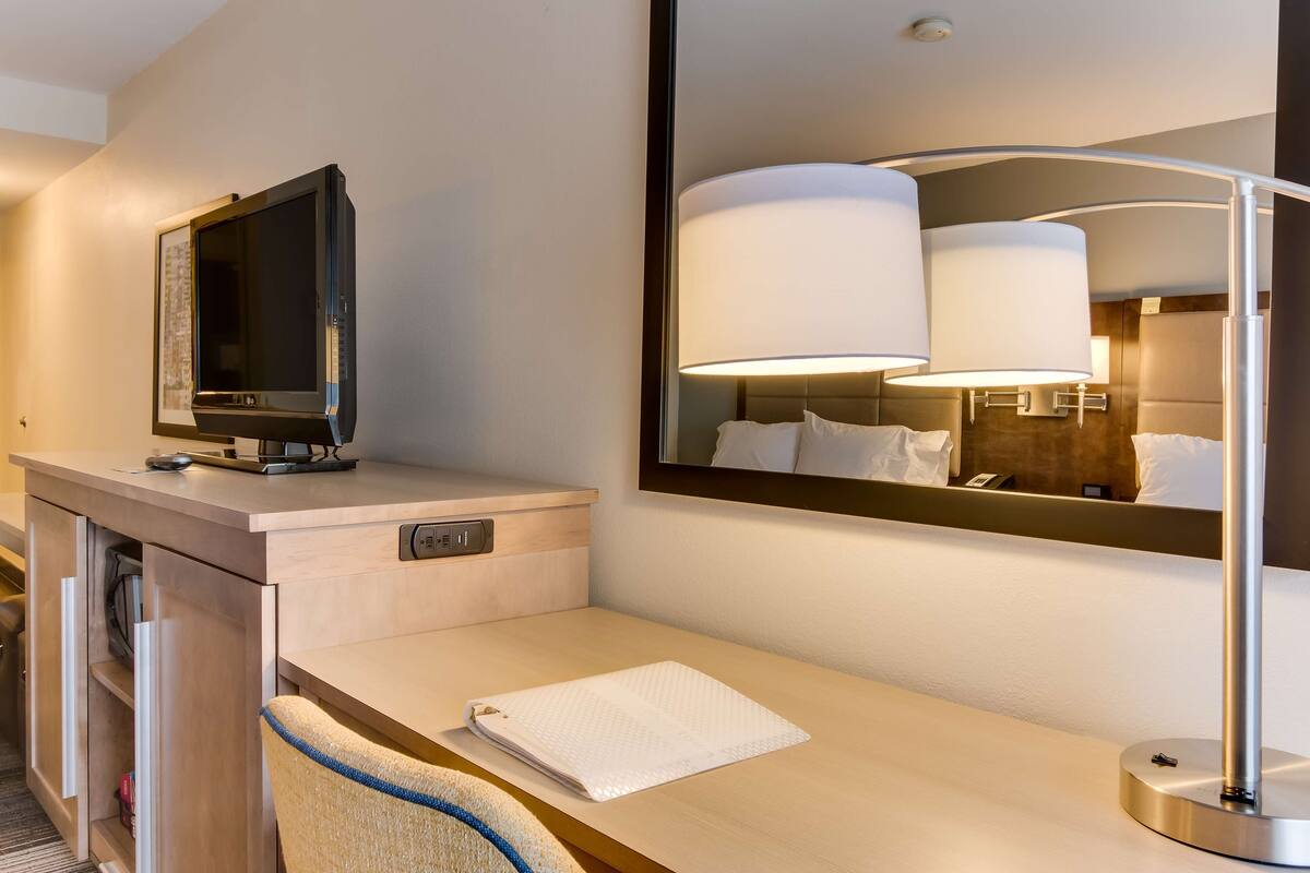 Habitación de hotel con escritorio, televisor de pantalla plana, lámpara, espejo y toalla sobre el mostrador.