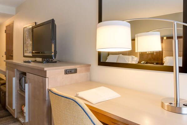 Quarto de hotel com televisão de tela plana sobre um console de madeira, um espelho grande refletindo uma cama com lençóis brancos e um abajur moderno.