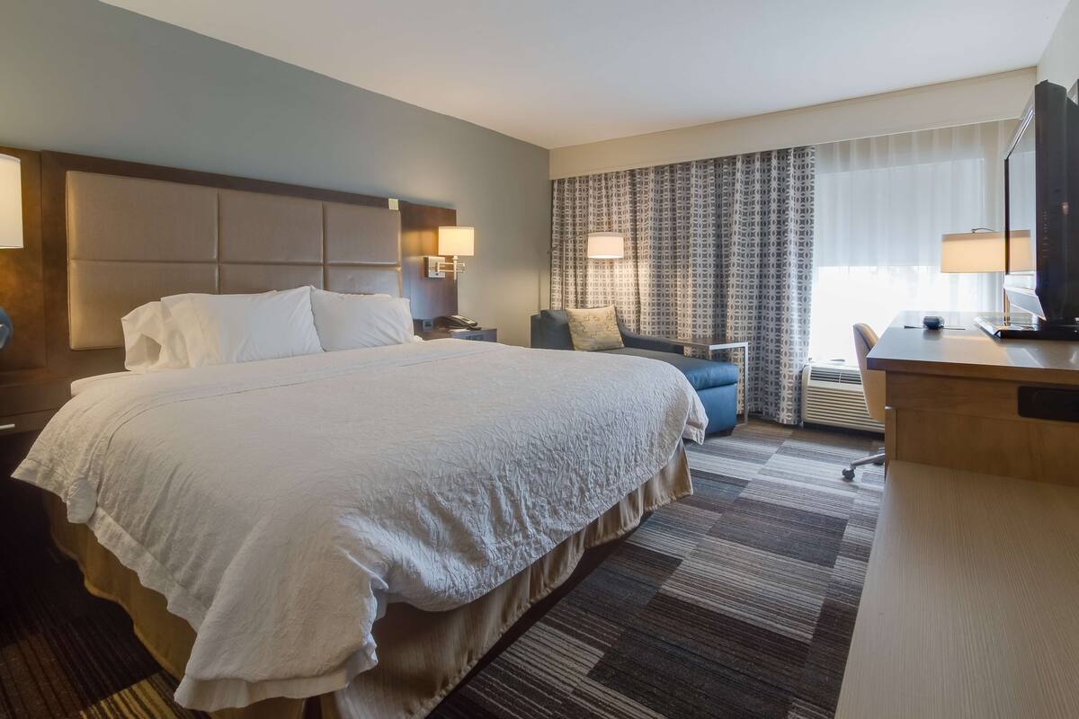 Una habitación de hotel con una cama grande, un escritorio, una silla, un televisor y una ventana con cortinas.