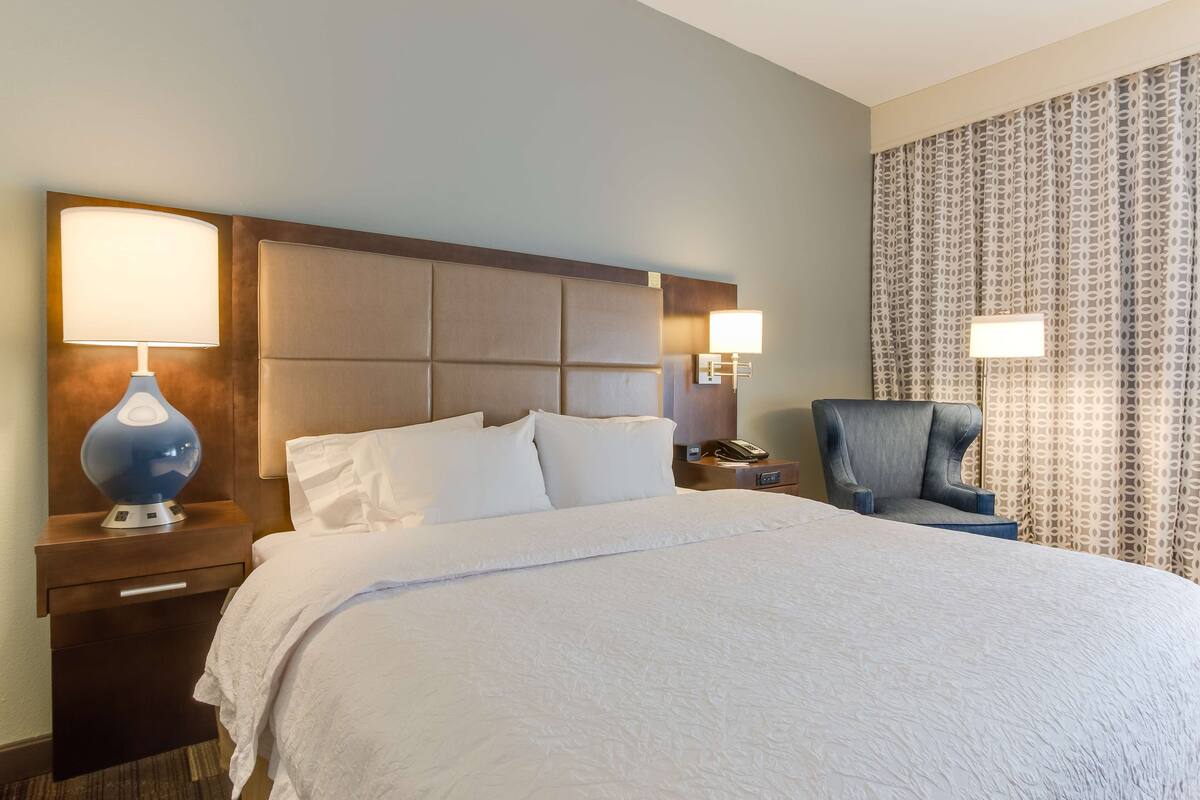 Habitación de hotel con una cama grande, una mesita de noche con lámpara, una silla y cortina estampada.