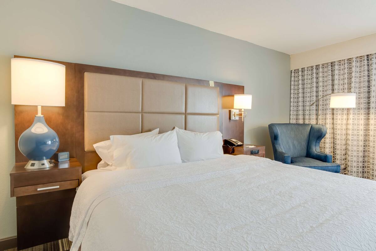 Habitación de hotel con una cama grande, una mesita de noche con lámpara, una silla y cortina estampada.