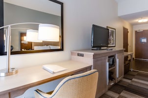 A hotel room with a desk, a chair, a television, a mirror, and a door.