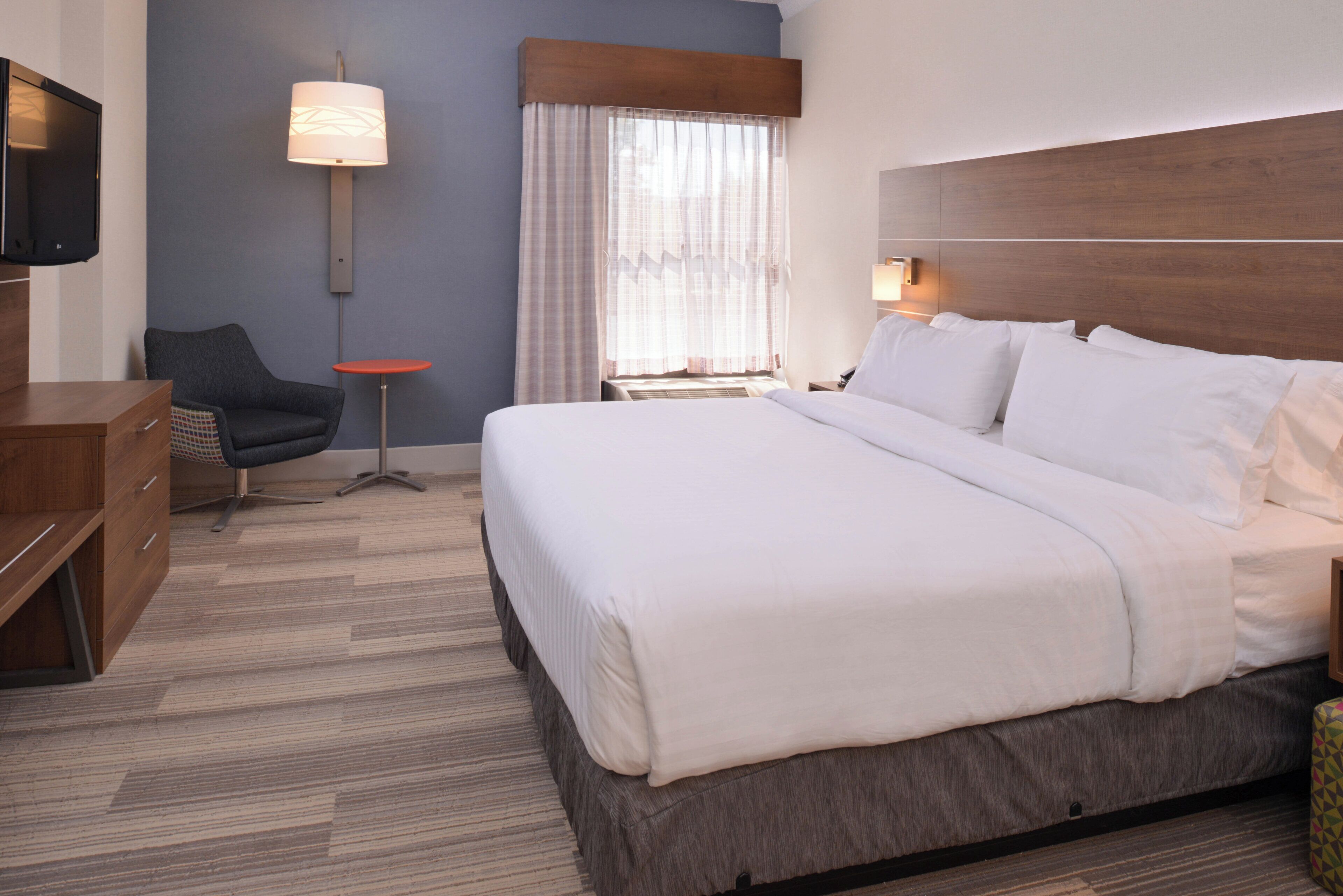 A hotel room with a bed, a TV, a chair, a small table, and a lamp.