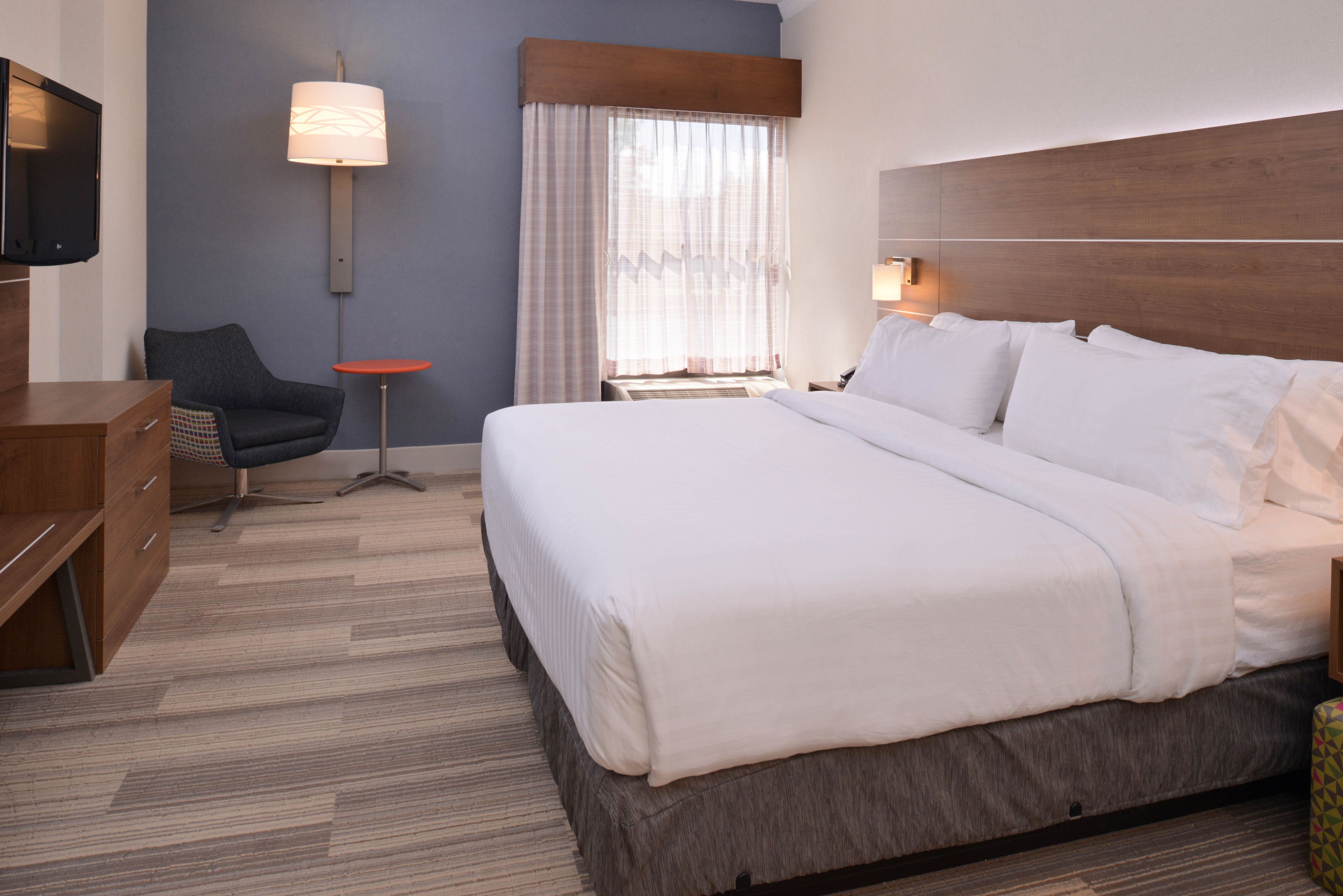 A hotel room with a large bed, a desk, a chair, a TV, a lamp, and a window with curtains.