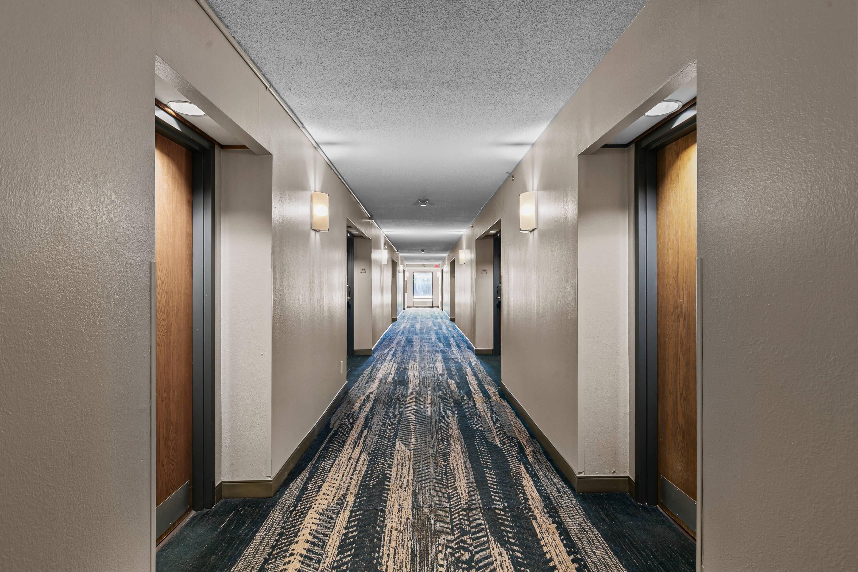A hotel hallway with carpeted floors, doors on both sides, and wall-mounted lighting.