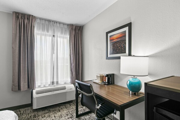 Habitación de hotel con escritorio, silla, aire acondicionado, cafetera, un cuadro en la pared y ventana con cortinas.