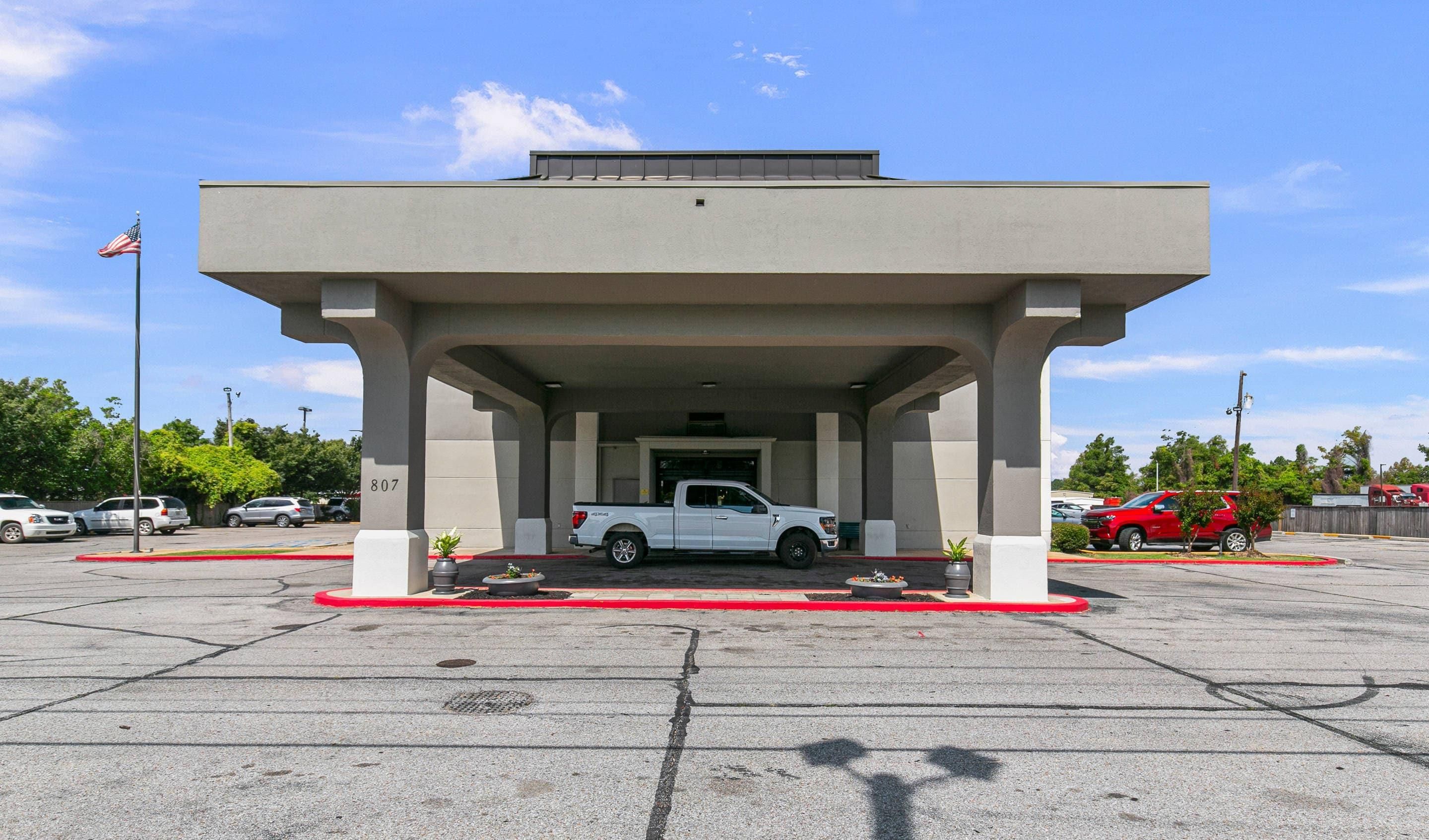 A building with a covered parking area, a white pickup truck parked inside, and a red car parked outside.
