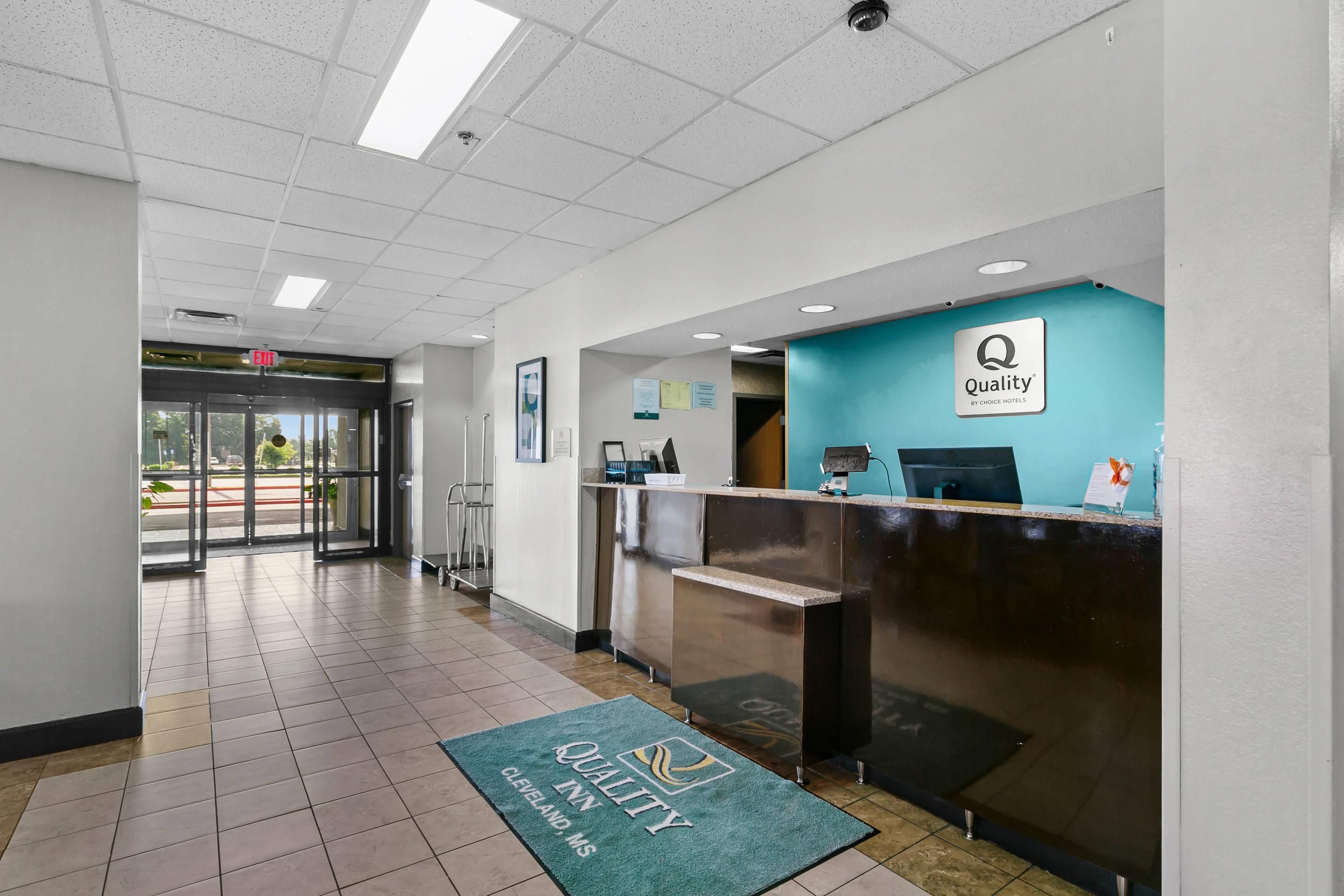 A hotel reception area with a reception desk, a blue wall with the hotel logo, a green carpet with the hotel name, and a view of the outside through the glass doors.
