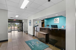 A hotel reception area with a reception desk, a blue wall with the hotel logo, a green carpet with the hotel name, and a view of the outside through the glass doors. - Quality Inn Cleveland, MS (Cleveland)