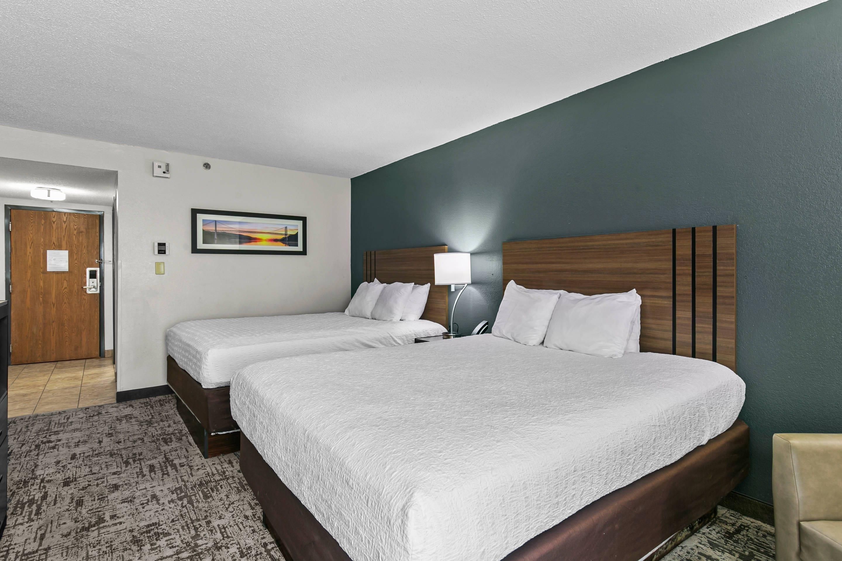 A hotel room with two beds, a nightstand, a lamp, a painting, and a door.