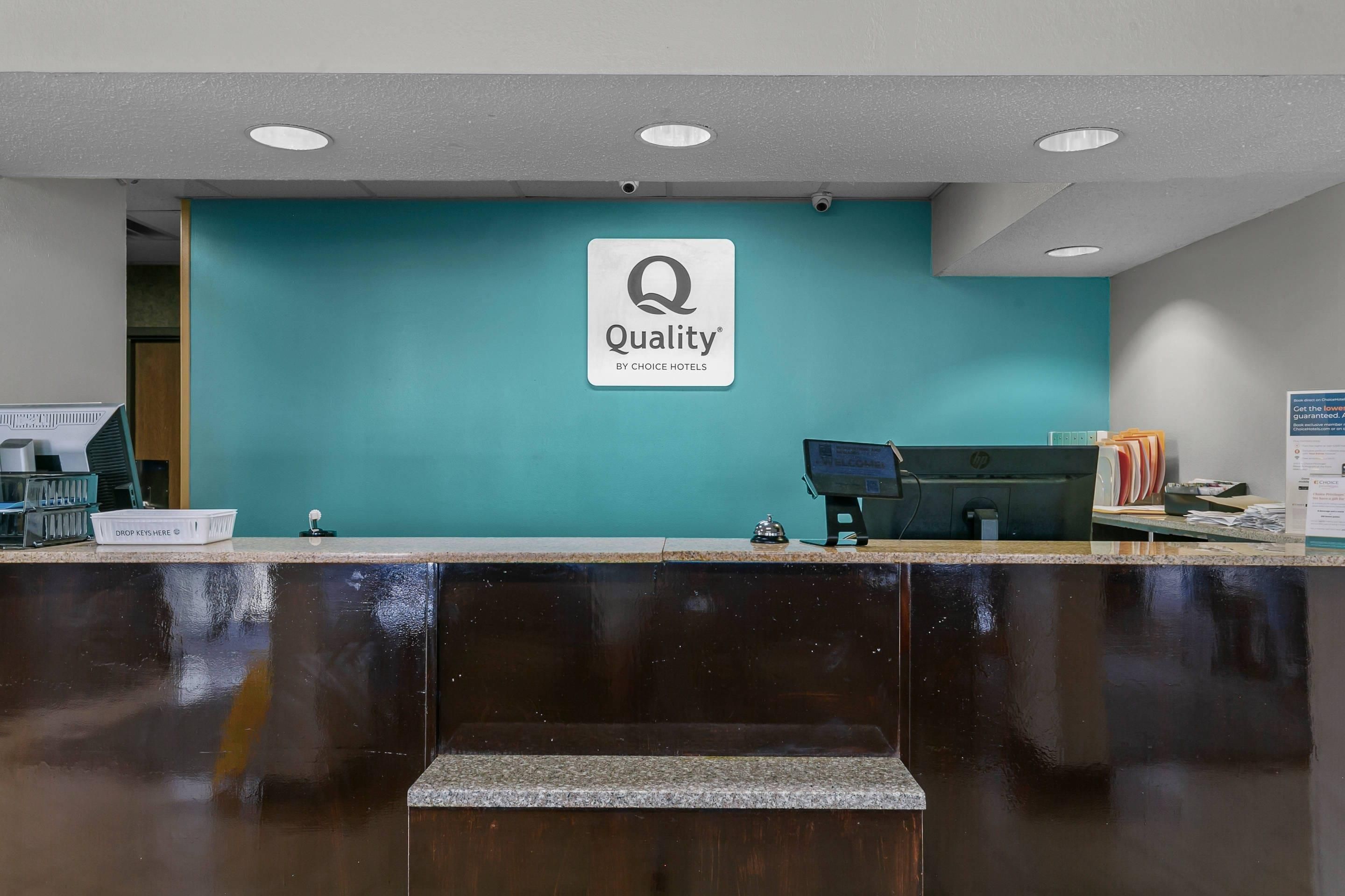 A hotel reception desk with a teal wall, a sign reading 'Quality by Choice Hotels', a computer monitor, and a cash register.