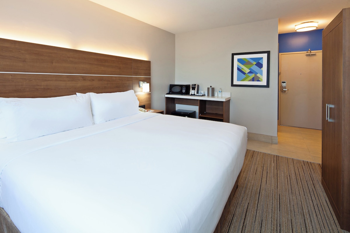 Une chambre d’hôtel avec un grand lit, une table de chevet avec une lampe, un micro-ondes, une petite étagère avec des articles de toilette et une porte donnant sur une autre pièce.