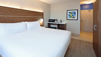 A hotel room with a large bed, a bedside table with a lamp, a microwave, a small shelf with toiletries, and a door leading to another room.