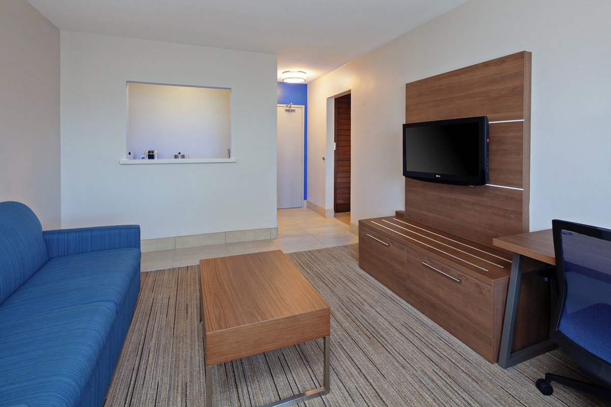 Une chambre d’hôtel moderne avec un canapé bleu, une table basse en bois, un téléviseur fixé sur un panneau en bois et un bureau avec une chaise.