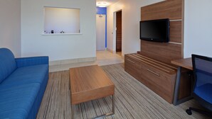 A modern hotel room with a blue sofa, a wooden desk, a flat-screen TV mounted on a wooden panel, and a small shelf with toiletries. - Holiday Inn Express Hotel & Suites San Diego-Escondido by IHG (Escondido)