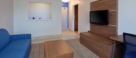 A modern hotel room with a blue sofa, a wooden desk, a flat-screen TV mounted on a wooden panel, and a small shelf with toiletries.