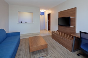 Et moderne hotelværelse med en blå sofa, et træskrivebord, et fladskærms-tv monteret på en træpanel og en lille hylde med toiletartikler.