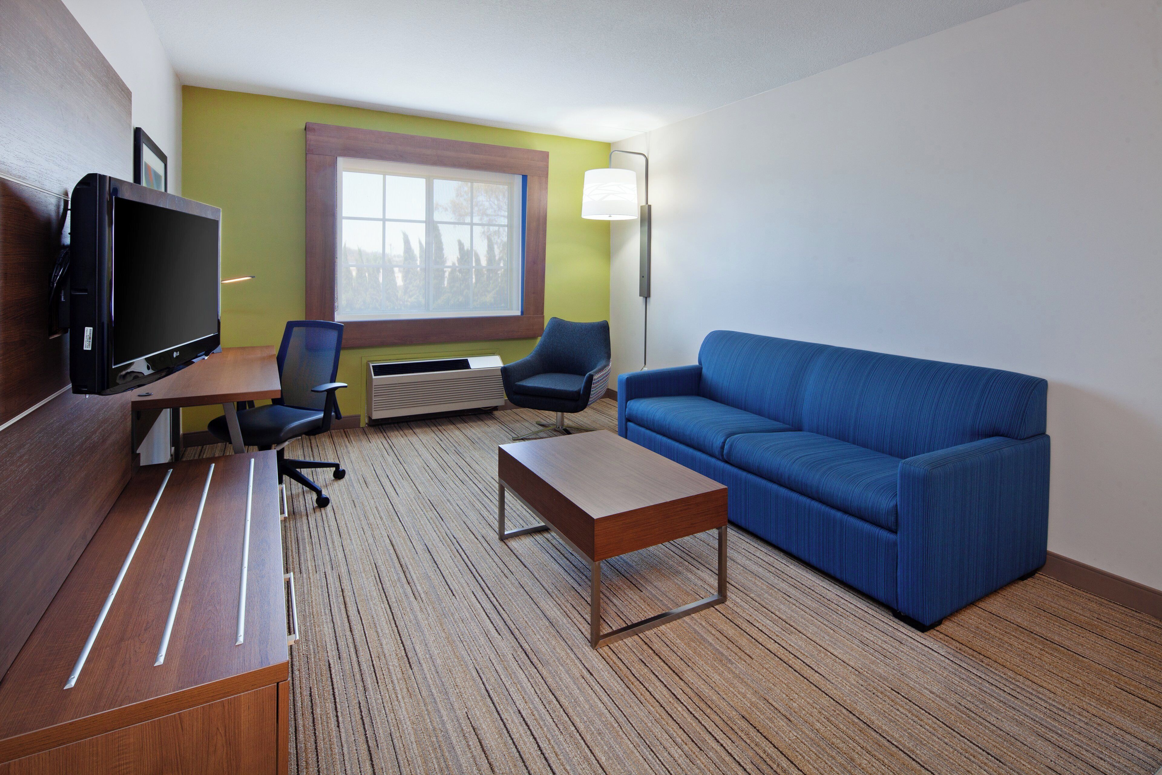 A hotel room with a flat-screen TV, a sofa, a coffee table, two chairs, a window, and a wall-mounted lamp.