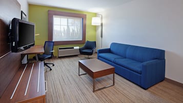 Quarto de hotel com televisão de tela plana, sofá, mesa de centro, duas cadeiras, janela e luminária fixada na parede.
