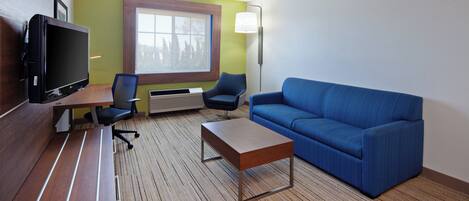 A hotel room with a flat-screen TV, a sofa, a coffee table, two chairs, a window, and a wall-mounted lamp.