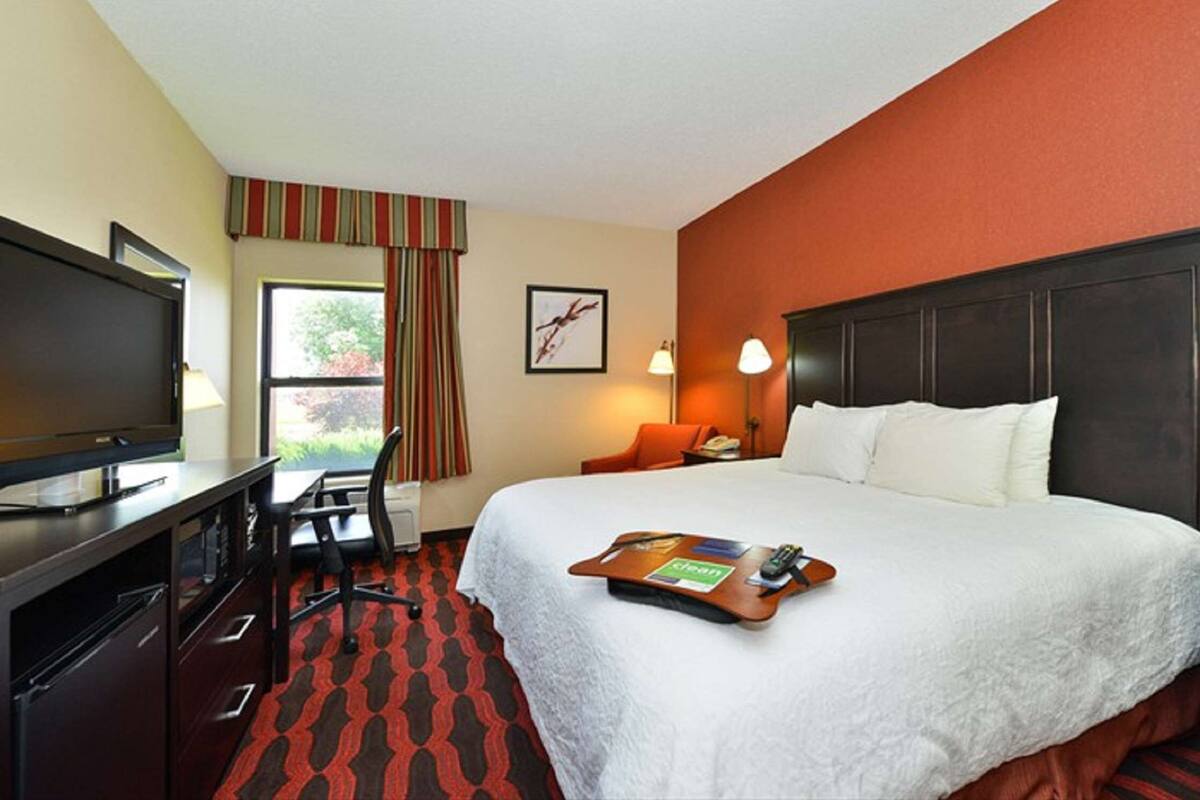 A hotel room with a bed, a desk with a TV, a chair, a window with curtains, a framed picture on the wall, and a bedside lamp.