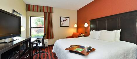 A hotel room with a bed, a desk with a TV, a chair, a window with curtains, a framed picture on the wall, and a bedside lamp.