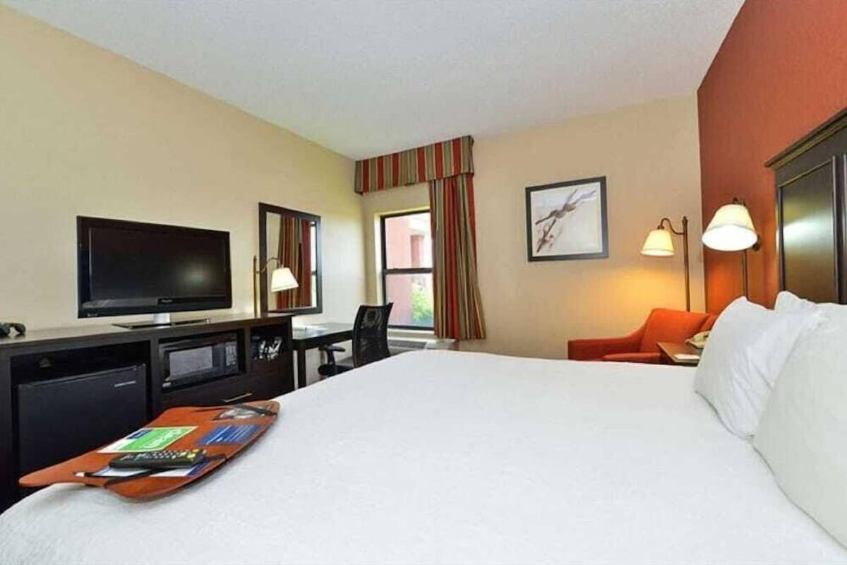 A hotel room with a bed, a desk with a TV, a chair, a lamp, and a window with curtains.