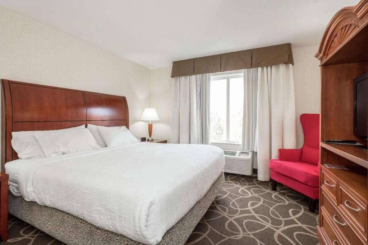 A hotel room with a large bed, a television, a red chair, and a window with curtains.