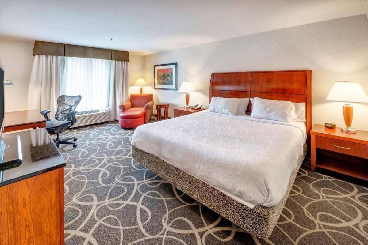 A hotel room with a large bed, a desk, a chair, a small table, and a window with curtains.